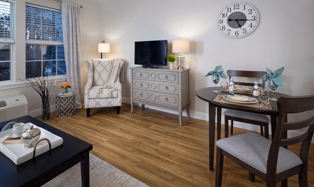 A cozy living and dining area with a patterned armchair next to a window with curtains, a small side table with flowers, a dresser with a TV and two lamps on top, and a round dining table set for two with plates, glasses, and napkins. A large wall clock is mounted above the dining table.