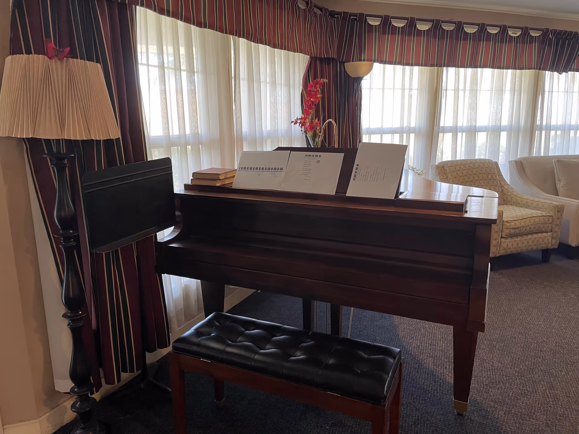 A cozy living room area with a dark wooden piano and a matching cushioned bench in front of large windows with sheer white curtains and striped valances. On the piano are some books, sheet music, and a vase with red flowers. To the right, there are two upholstered chairs, one patterned and one plain light-colored. A tall floor lamp with a pleated shade stands to the left of the piano.