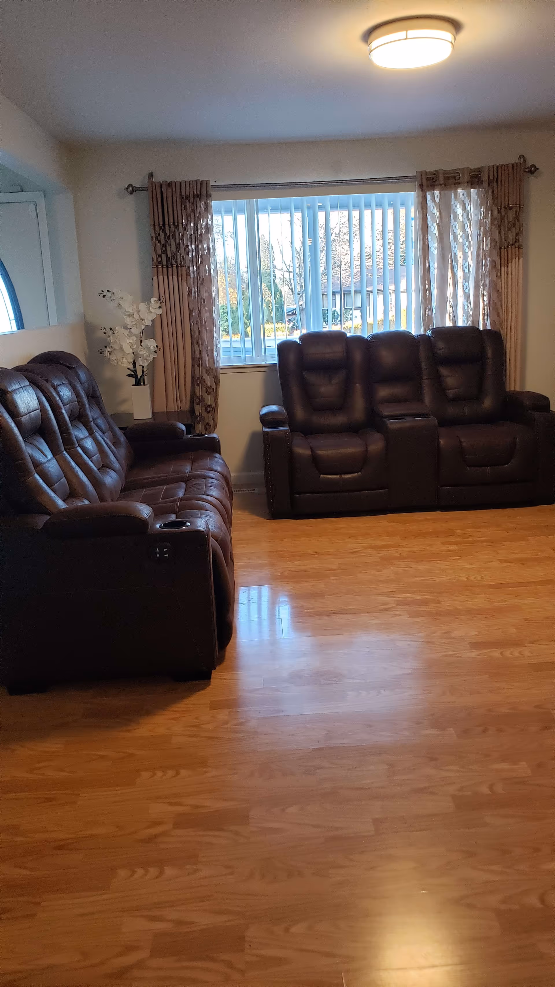 A living room with two dark brown leather recliner sofas facing each other. There is a large window with vertical blinds and patterned curtains behind one sofa. A white vase with white flowers is placed on a small table near the window. The floor is wooden, and a ceiling light fixture is visible.