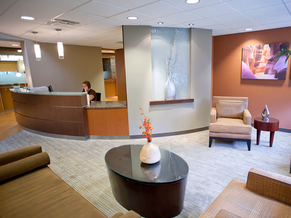 Reception area of a healthcare facility with a curved wooden reception desk where a person is seated and talking on the phone. The room has modern decor with a beige armchair, a small round wooden side table with a decorative object, a dark oval coffee table with a vase containing orange flowers, and a large wall art piece. The floor is carpeted and the walls are painted in neutral tones.