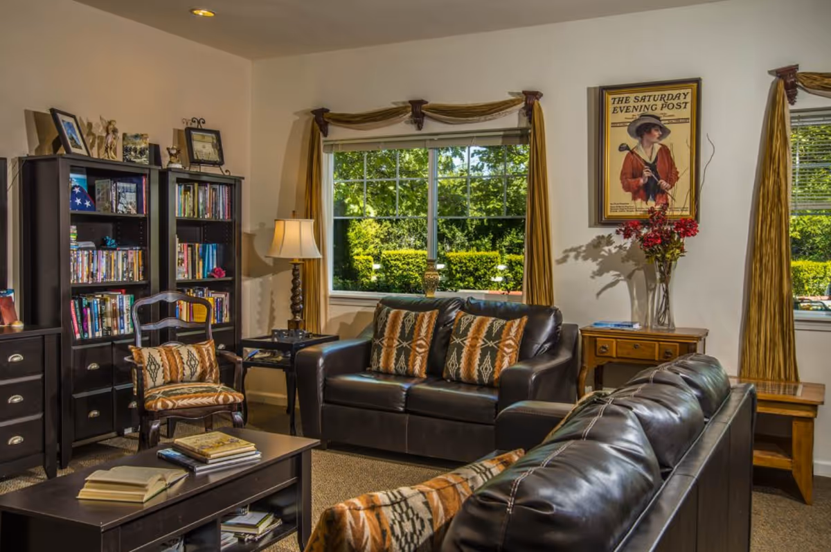 A cozy living room with two black leather sofas adorned with patterned cushions, a wooden coffee table with books, a wooden chair with a matching cushion, a floor lamp, two black bookshelves filled with books and decorative items, and a window with golden curtains showing green bushes outside. A framed vintage poster titled 'The Saturday Evening Post' and a vase with red flowers are on a wooden side table.