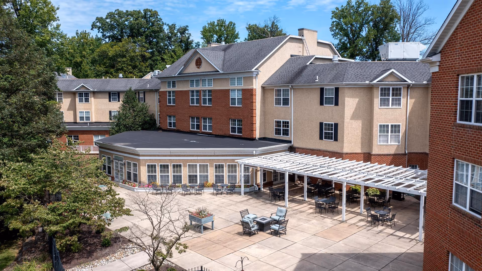 Courtyard at Rockland Place with a pergola, patio tables and chairs in front of a multi-story brick-and-stucco building surrounded by trees.