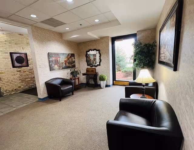 A cozy sitting area in an assisted living facility with three black leather armchairs arranged around a small wooden side table with a lamp. The walls are decorated with framed artwork and a decorative mirror. There is a large window letting in natural light and a potted plant near the window. The floor is carpeted and the ceiling has recessed lighting.
