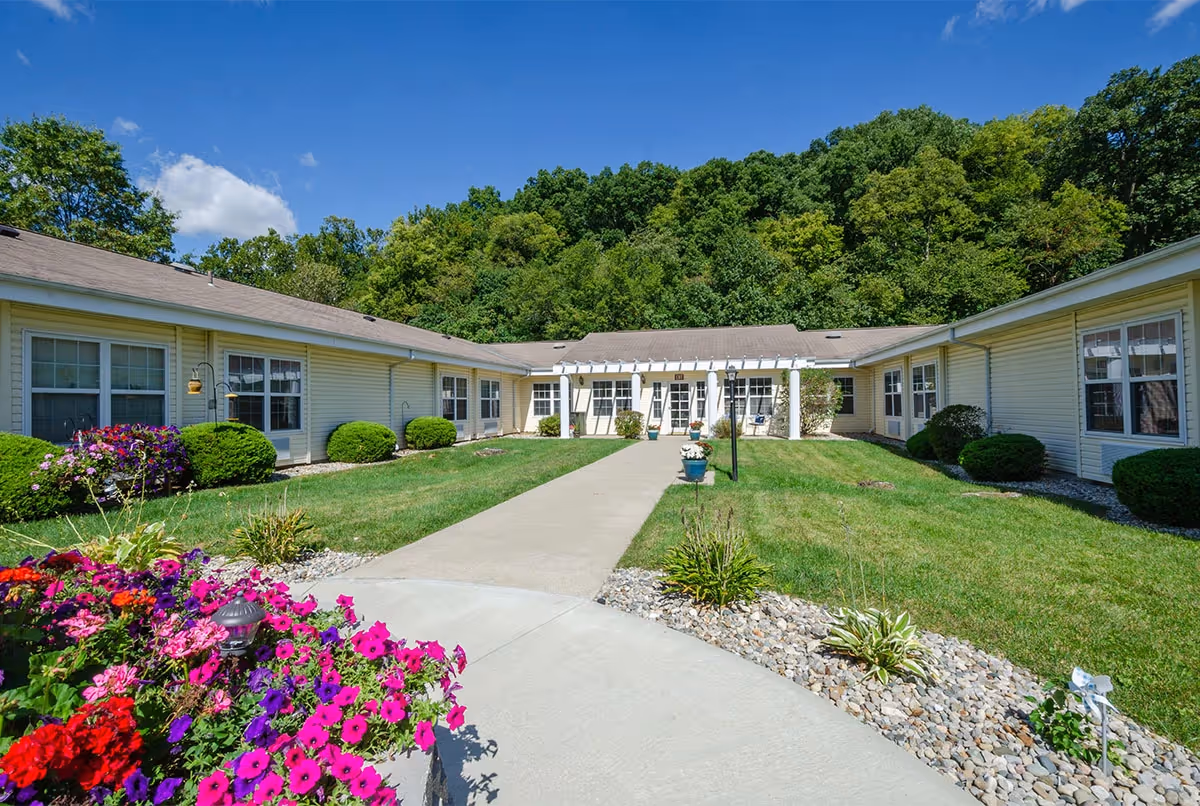 Courtyard entrance of a single-story senior living building with a central walkway, lawns, flowerbeds and wooded hills behind.
