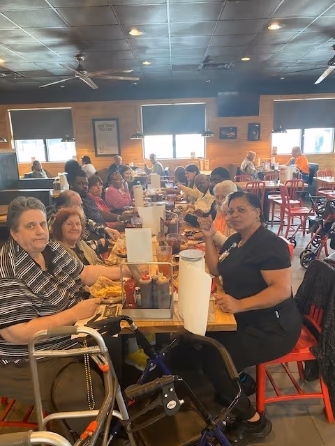 A group of elderly people and a caregiver sitting around a long table in a dining area, enjoying a meal together. The room has wooden walls, large windows with blinds, ceiling fans, and red chairs. Some people have walkers nearby.