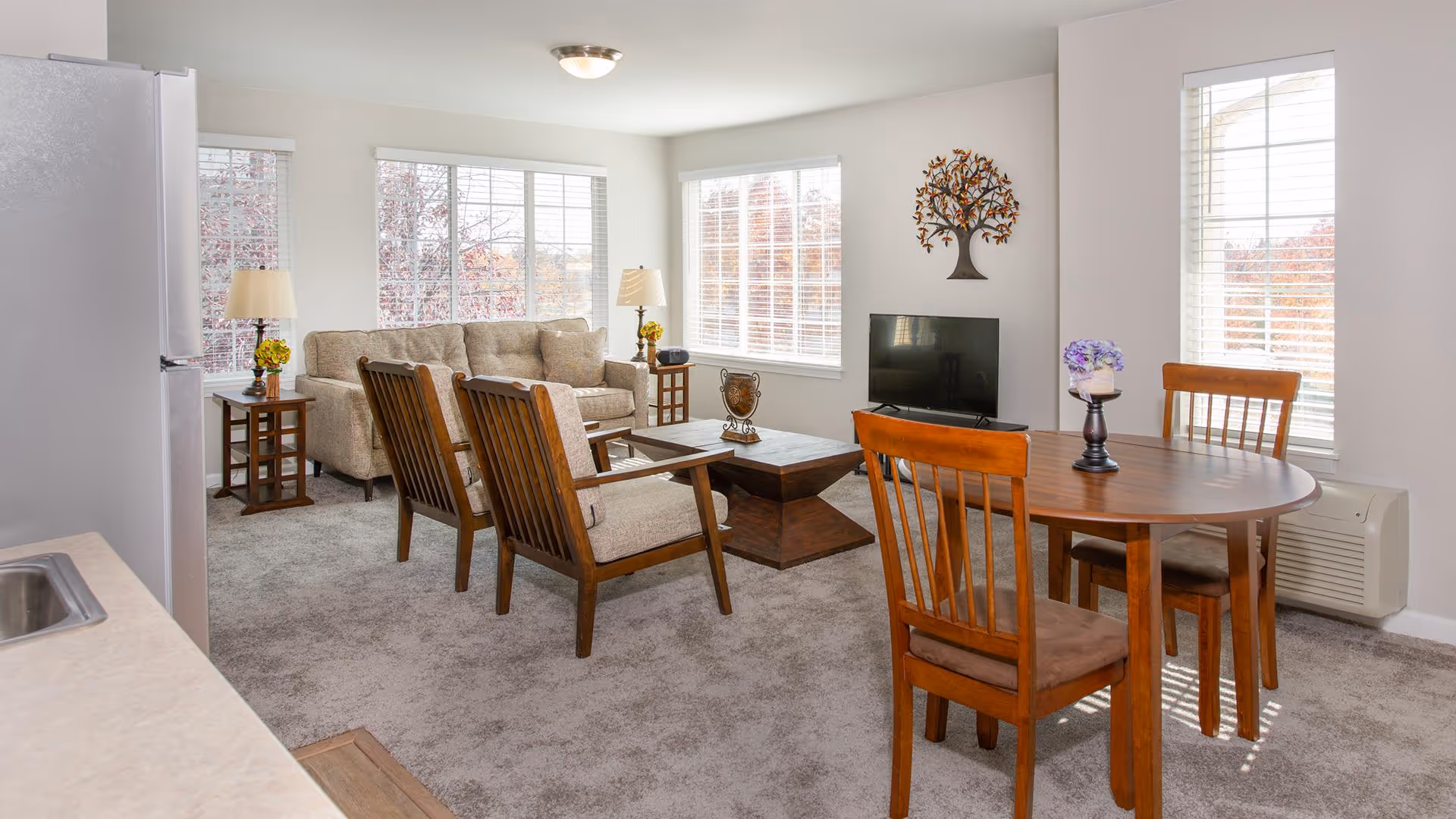 A bright living room and dining area with large windows letting in natural light. The living room features a beige sofa, two wooden armchairs with cushions, a wooden coffee table, and two side tables with lamps and flowers. The dining area has a round wooden table with four chairs and a small vase with purple flowers. A flat-screen TV is mounted on a stand against the wall, and a decorative metal tree wall art is above it. The room has light-colored walls and carpeted flooring.