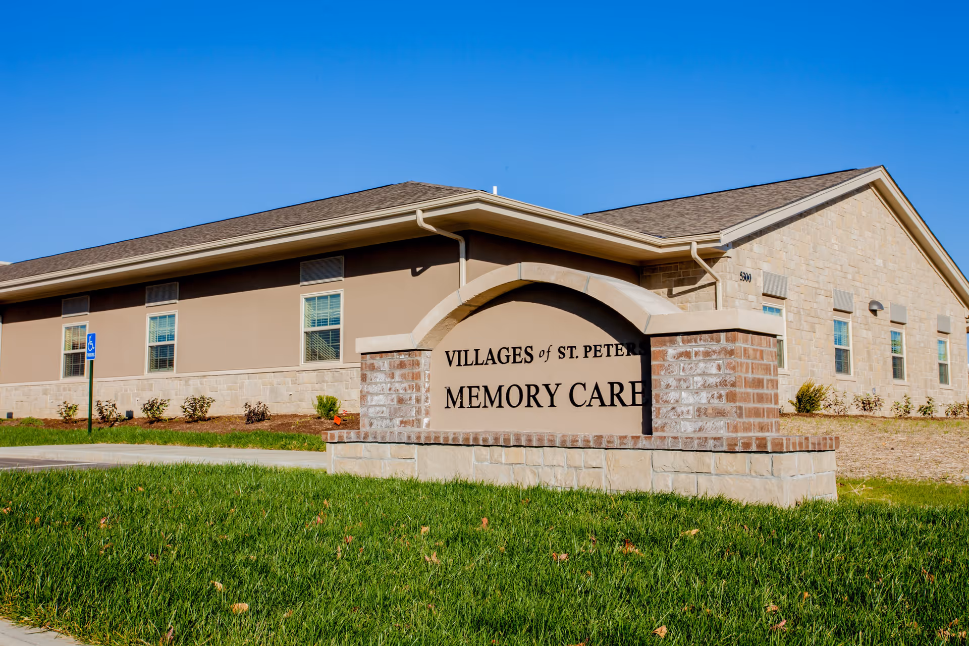 Exterior view of a single-story memory care facility building with beige and stone walls under a clear blue sky. In front of the building is a large stone and brick sign that reads 'Villages of St. Peters Memory Care'. There is a small grassy area and a sidewalk in the foreground.