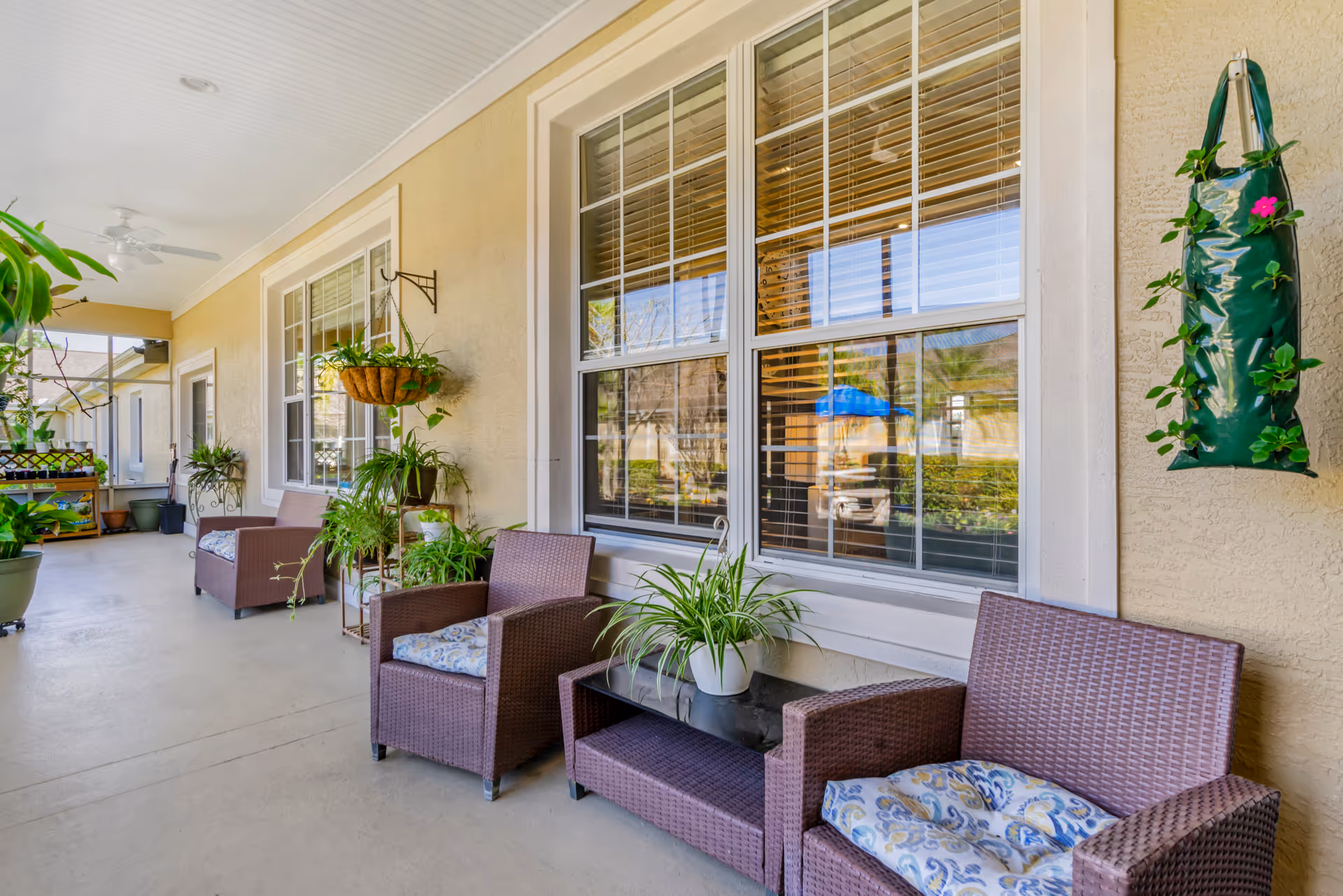 Covered outdoor porch with wicker chairs, potted plants, hanging planters, and windows along a beige building.