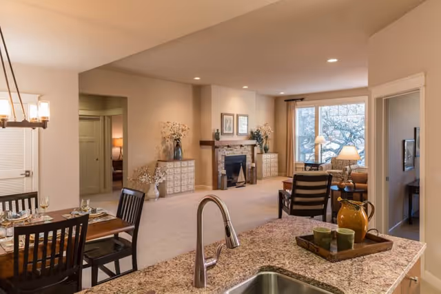 View of an open-concept living space in a senior living facility showing a kitchen island with a sink and a tray with a pitcher and cups, a dining area with a table set for four, and a living room with chairs, a fireplace, and large windows letting in natural light.