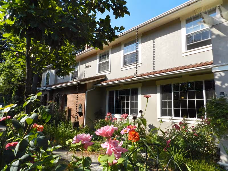 Exterior view of a two-story senior living facility building with beige and brick walls, multiple windows, and a garden with blooming flowers and greenery in the foreground.