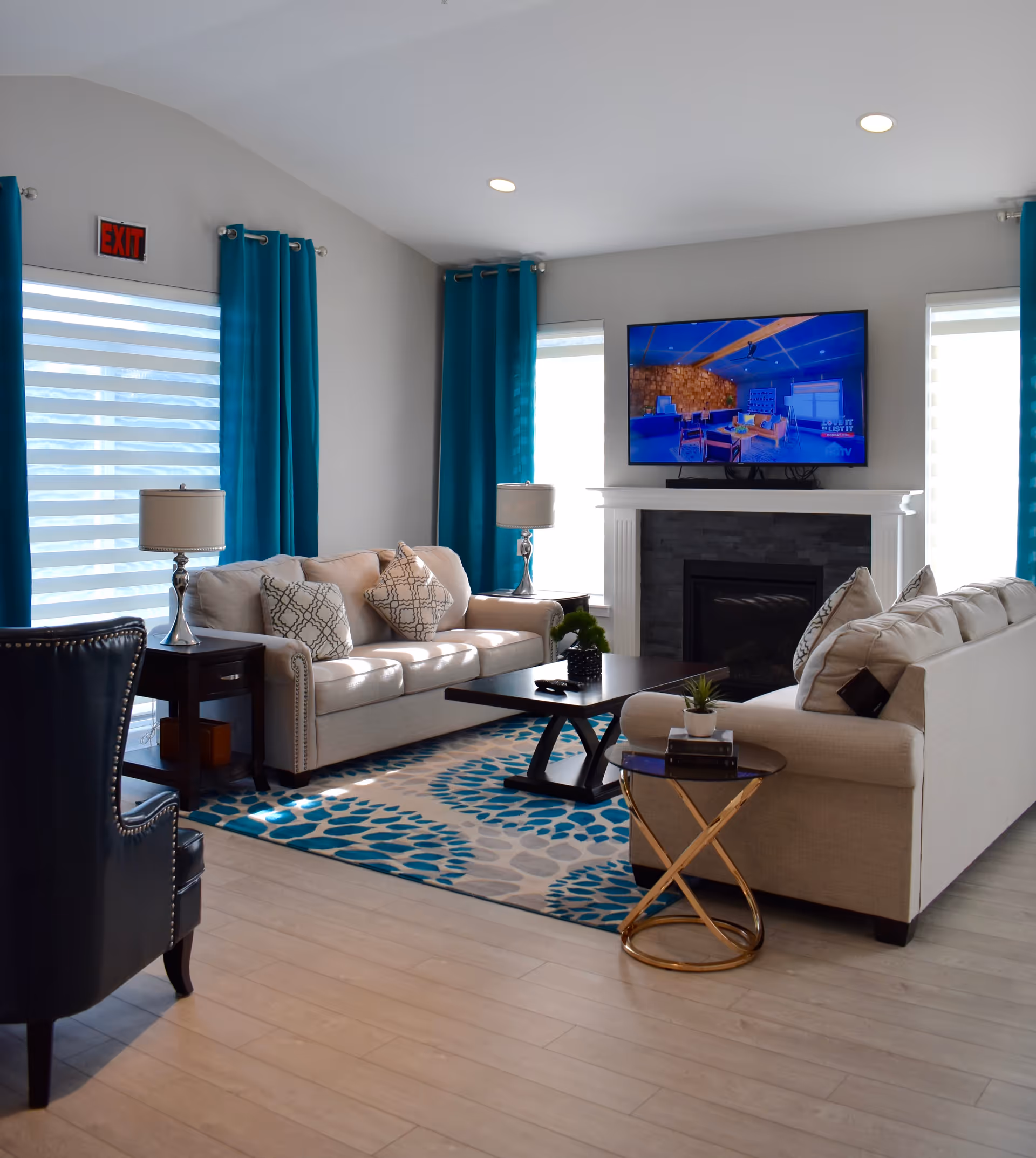 A bright and modern living room with two beige sofas adorned with patterned cushions, a dark wooden coffee table, and a small round side table with books and a plant. The room features large windows with teal curtains, a fireplace with a mounted flat-screen TV above it, and light wood flooring.