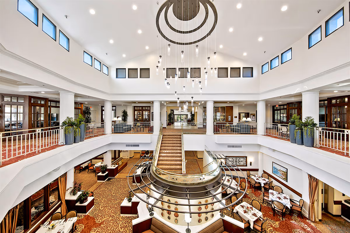 Two-story bright atrium with a central staircase, hanging chandelier, balconies, and dining tables on the lower level.