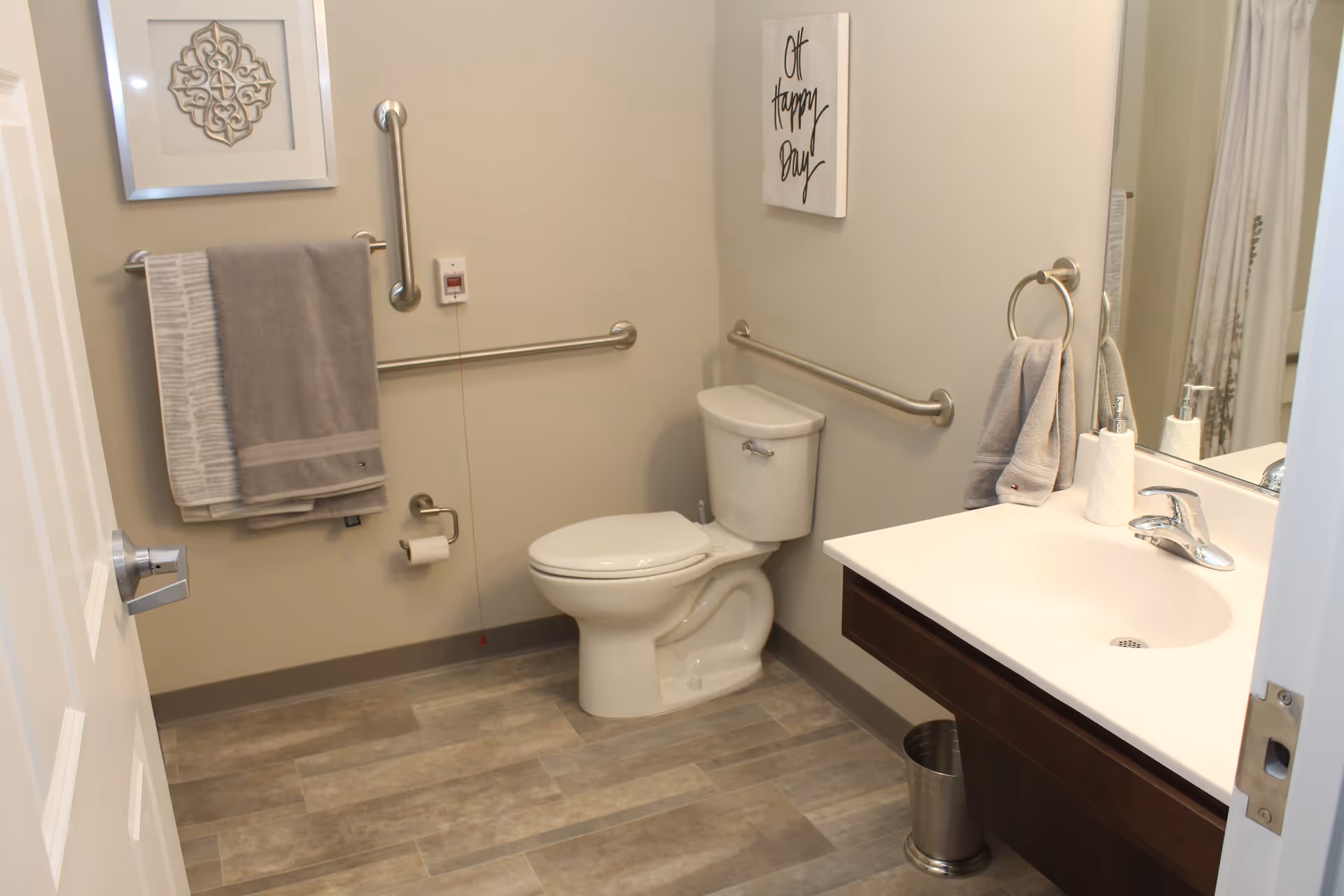 A clean and accessible bathroom with a white toilet, grab bars on the walls, a sink with a faucet, a mirror, and towels hanging on racks. The floor has a wood-like tile pattern, and there is wall art with the phrase 'Oh happy day'.