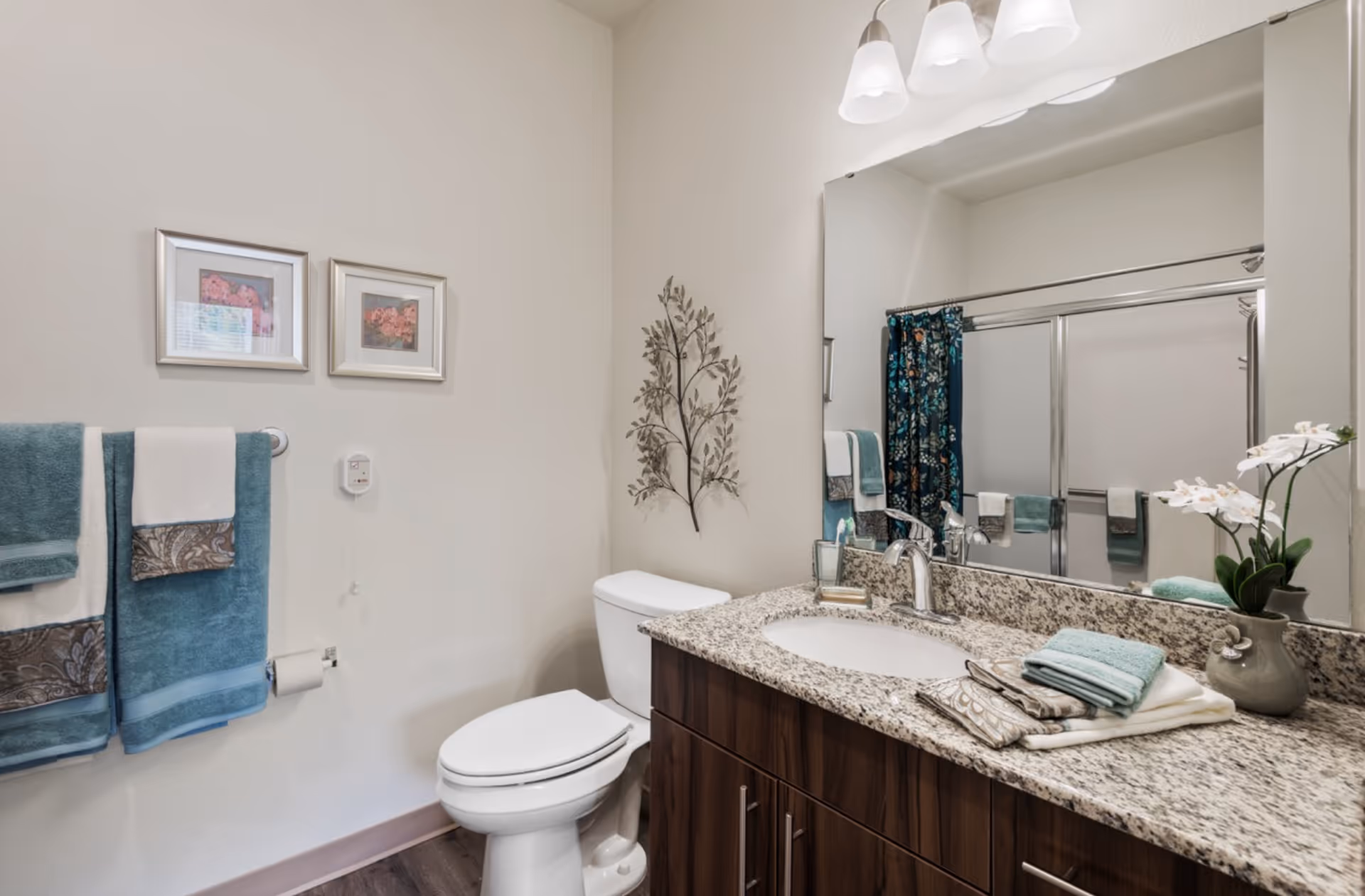 A clean and well-decorated bathroom featuring a white toilet, a granite countertop with a sink, and a large mirror above the sink. The countertop holds neatly folded towels and a small vase with white flowers. On the wall above the toilet is a decorative metal wall art piece. Two framed pictures hang on the adjacent wall above a towel rack with teal and white towels. A shower with sliding glass doors and a floral shower curtain is visible in the mirror's reflection. The bathroom has light-colored walls and dark wood cabinetry under the sink.