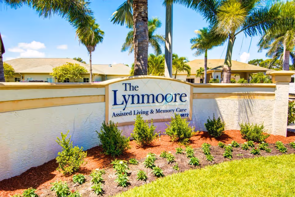 Entrance sign for The Lynmoore Assisted Living & Memory Care facility surrounded by landscaped plants and palm trees with residential buildings in the background under a blue sky.