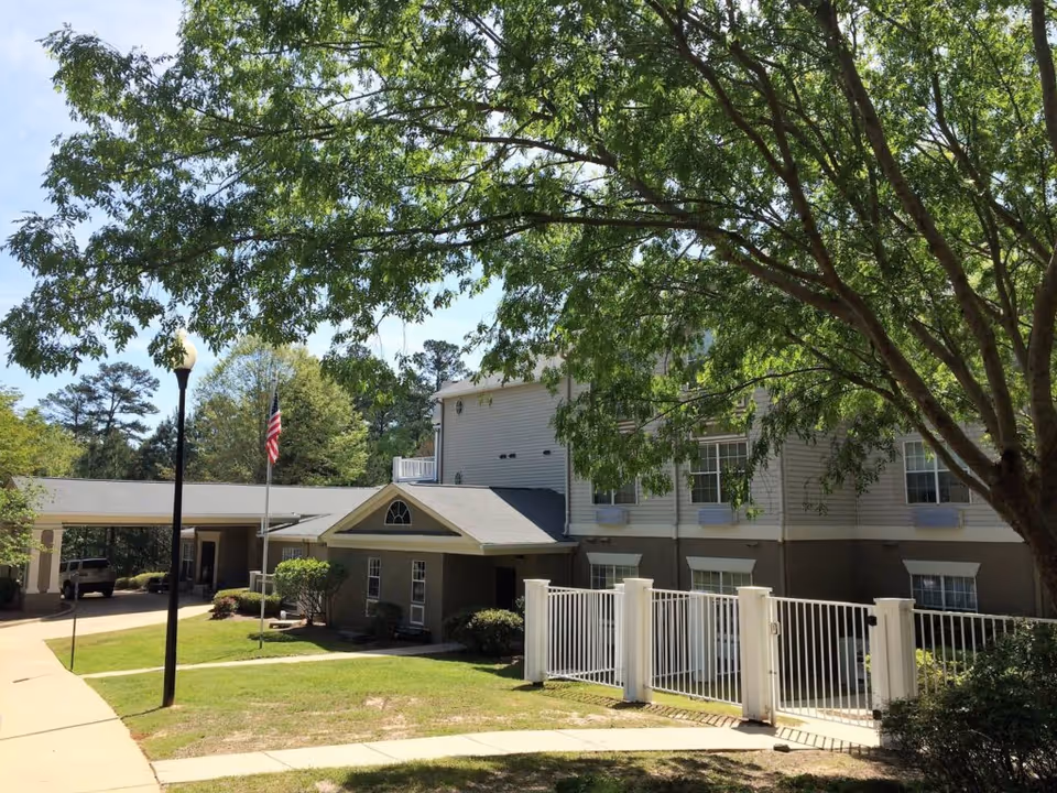Exterior of a two-story senior living building with a gated entrance, American flag, driveway, and large shade trees.