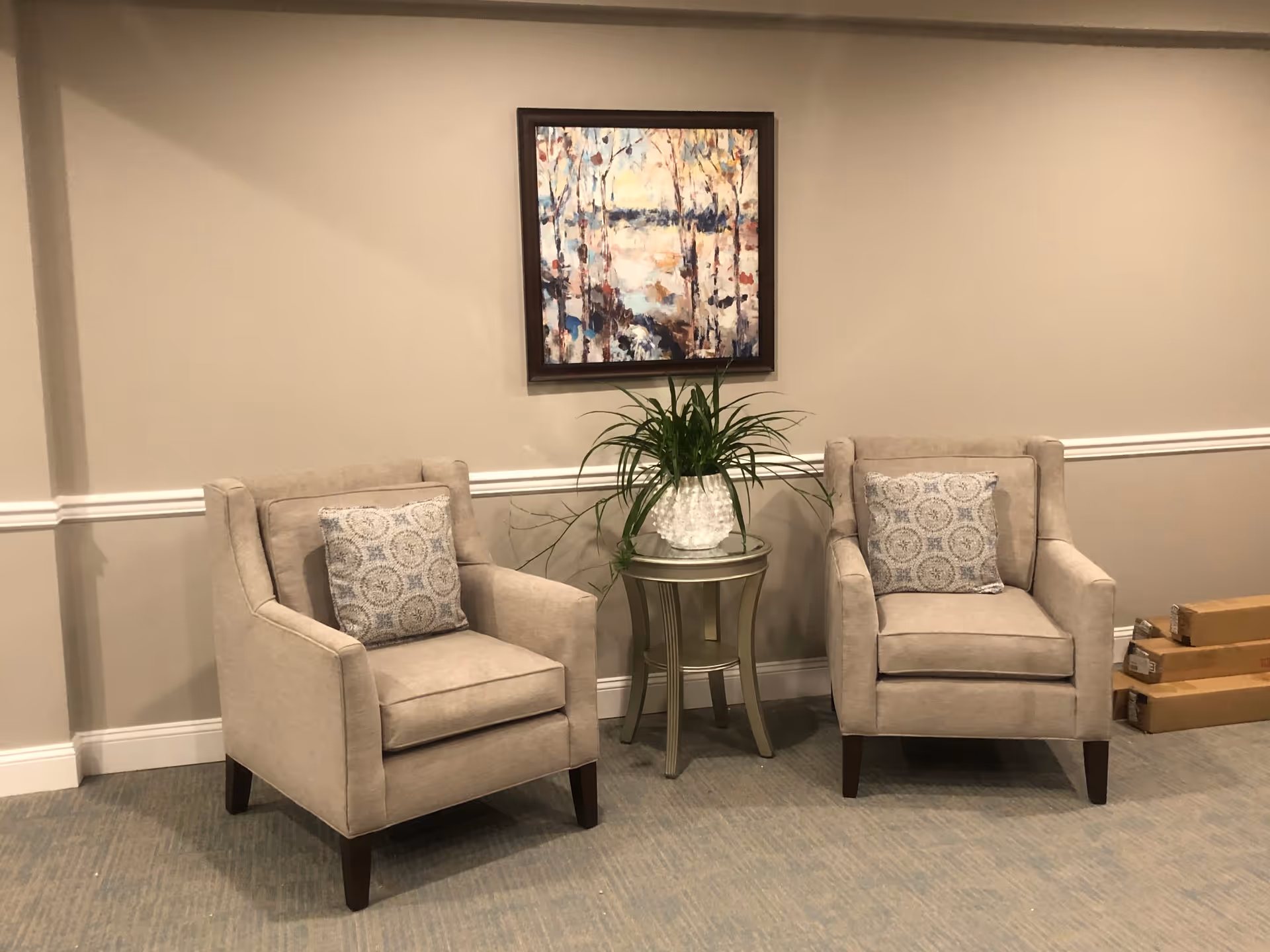 Two beige armchairs with patterned cushions are placed on either side of a small round table with a potted plant. Above the table hangs a framed painting of a nature scene with trees and water. The setting is indoors with beige walls and carpeted floor. There are three cardboard boxes stacked on the floor to the right.