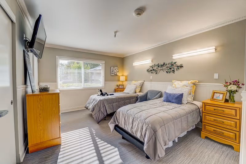 A bright and cozy bedroom with two single beds, each with gray bedding and decorative pillows. Between the beds is a wooden nightstand with a framed photo and a vase of flowers. A wooden dresser is positioned against the opposite wall beneath a mounted flat-screen TV. A large window with white blinds lets in natural light, and the walls are painted a soft beige with white wainscoting. The room has a calm and inviting atmosphere.