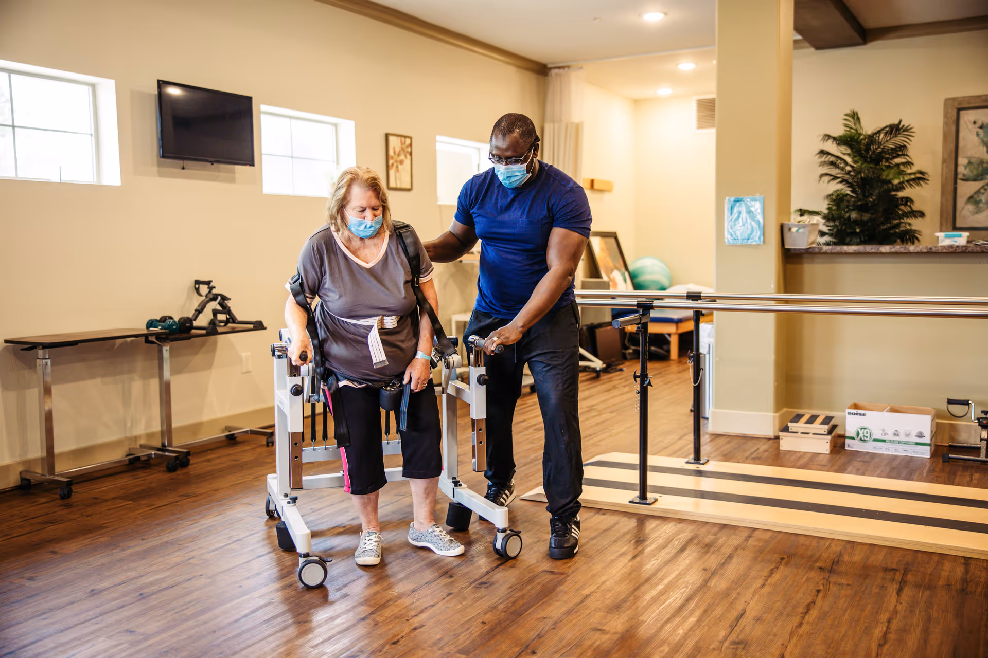 A woman using a walker is assisted by a man in a blue shirt and black pants in a rehabilitation room with wooden floors, parallel bars, and exercise equipment. Both individuals are wearing face masks.