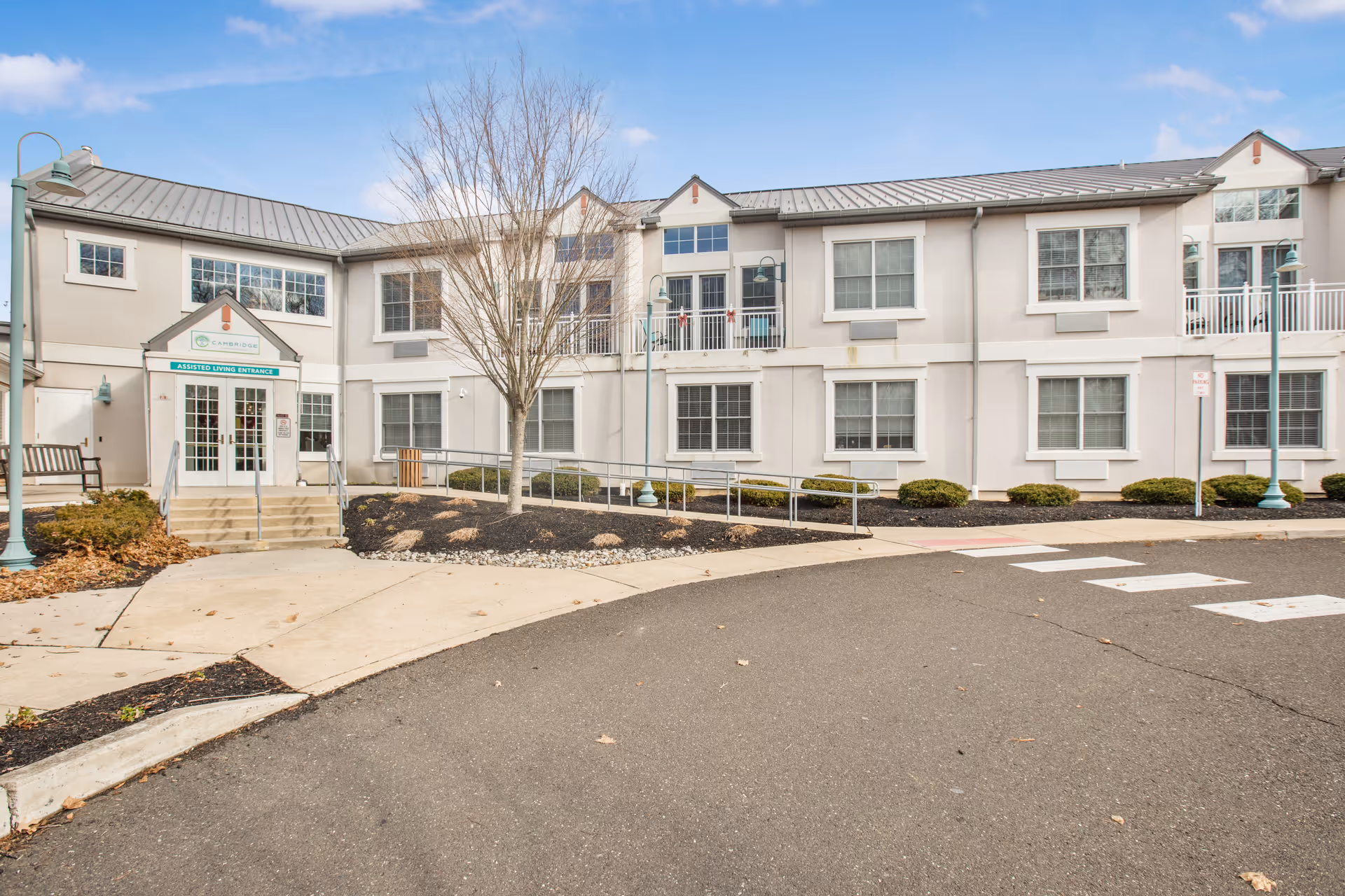Front exterior of a two-story senior living facility with a main entrance, ramp, bare tree, and driveway turnaround.