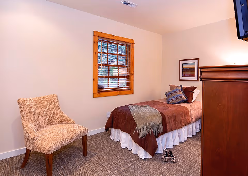 Cozy single bedroom with a twin bed, patterned throw pillows and blanket, an upholstered chair, wooden-window blinds, and a dresser.