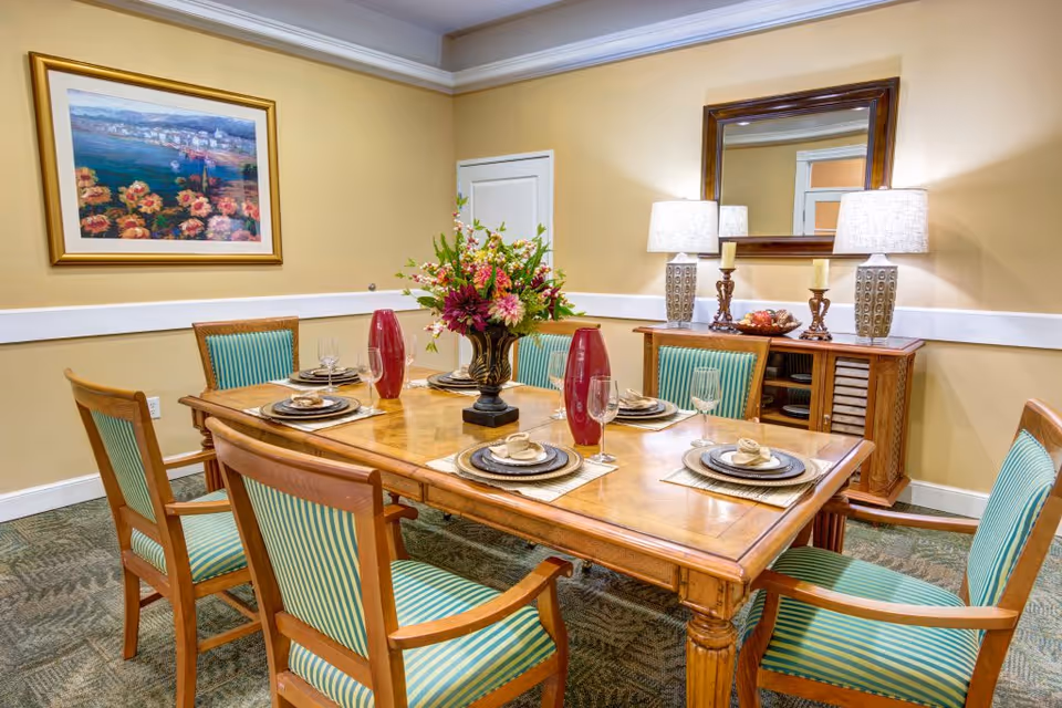 A dining room with a wooden table set for six people, featuring striped green and beige upholstered chairs. The table is decorated with a floral centerpiece, two red vases, and place settings with plates, napkins, and wine glasses. In the background, there is a wooden sideboard with two lamps, candles, and a bowl of fruit, along with a large mirror above it. A framed painting of flowers and a coastal scene hangs on the wall.