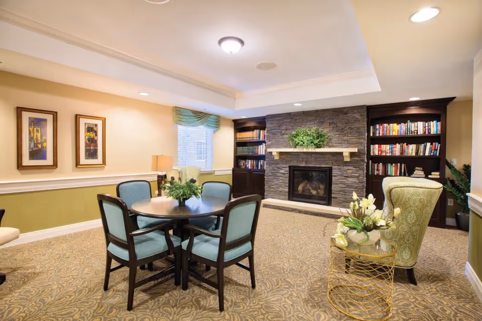 A cozy living room area with a round table surrounded by four chairs with blue cushions. There is a stone fireplace with a green plant on the mantel, flanked by built-in bookshelves filled with books. A green upholstered armchair sits next to a small round gold side table with a floral arrangement. The room has beige walls with two framed pictures, a window with a green valance, and recessed lighting in the ceiling.