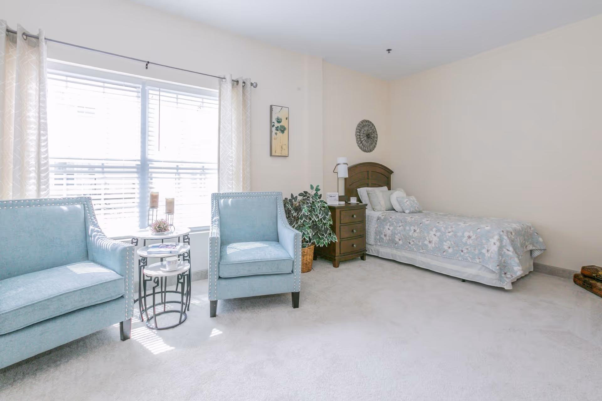 Bright furnished bedroom with a twin bed, nightstand and lamp, two blue armchairs by a large window, and decorative accents.