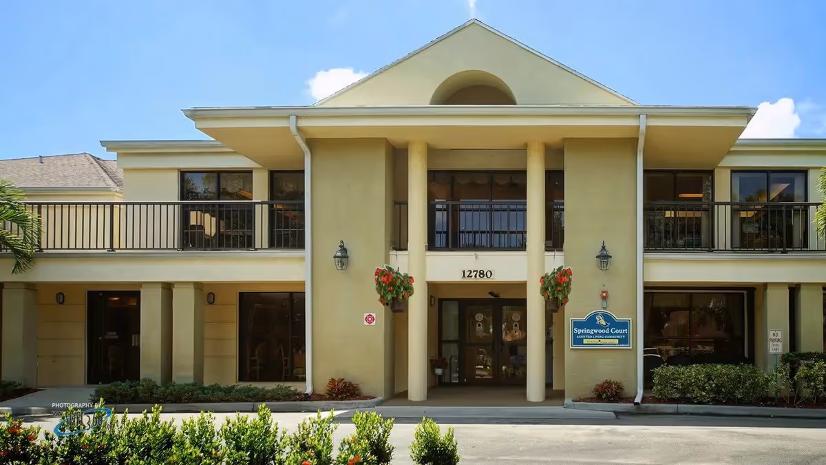 Front exterior view of a two-story building with beige walls, large windows, and a covered entrance supported by two columns. There are hanging flower pots on either side of the entrance and a sign that reads 'Springwood Court Assisted Living Community' near the door. The building number 12780 is displayed above the entrance.