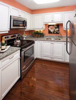 A modern kitchen with white cabinets, granite countertops, and stainless steel appliances including a stove, microwave, and refrigerator. The walls are painted a warm coral color, and there is a floral painting above the sink. The floor is a polished dark wood.
