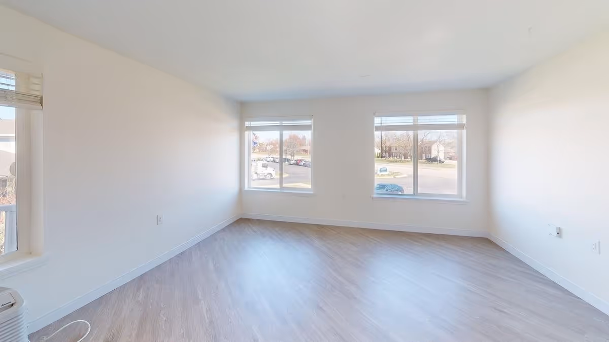 Empty room with light-colored walls and wood flooring, featuring three large windows with white blinds allowing natural light to enter. The room appears clean and spacious with electrical outlets visible on the walls.