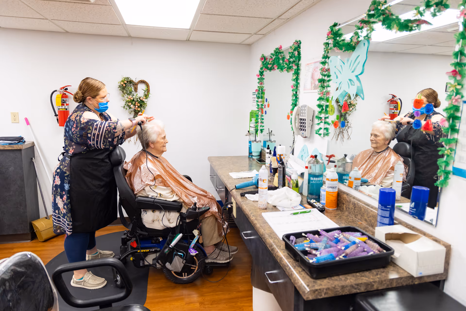 A masked hairstylist trims an elderly woman's hair in a salon area with a large mirror, counter full of hair products, and wall decorations.