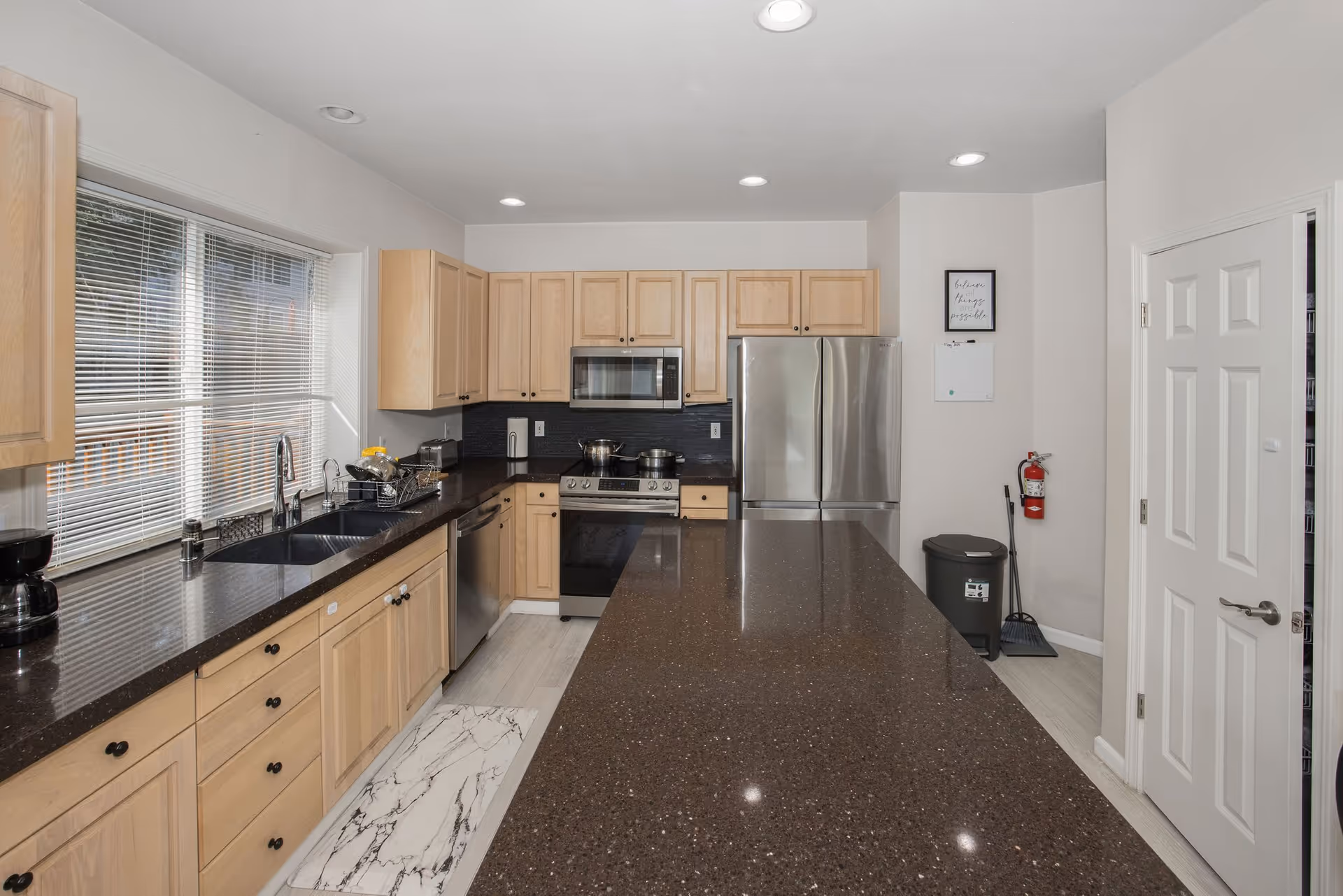 A modern kitchen with light wood cabinets, a large dark brown speckled countertop island, stainless steel appliances including a refrigerator, microwave, stove, and dishwasher. There is a double sink under a window with blinds, a coffee maker on the counter, and a fire extinguisher mounted on the wall near a white door. The floor has light-colored tiles and a marble-patterned rug in front of the sink.