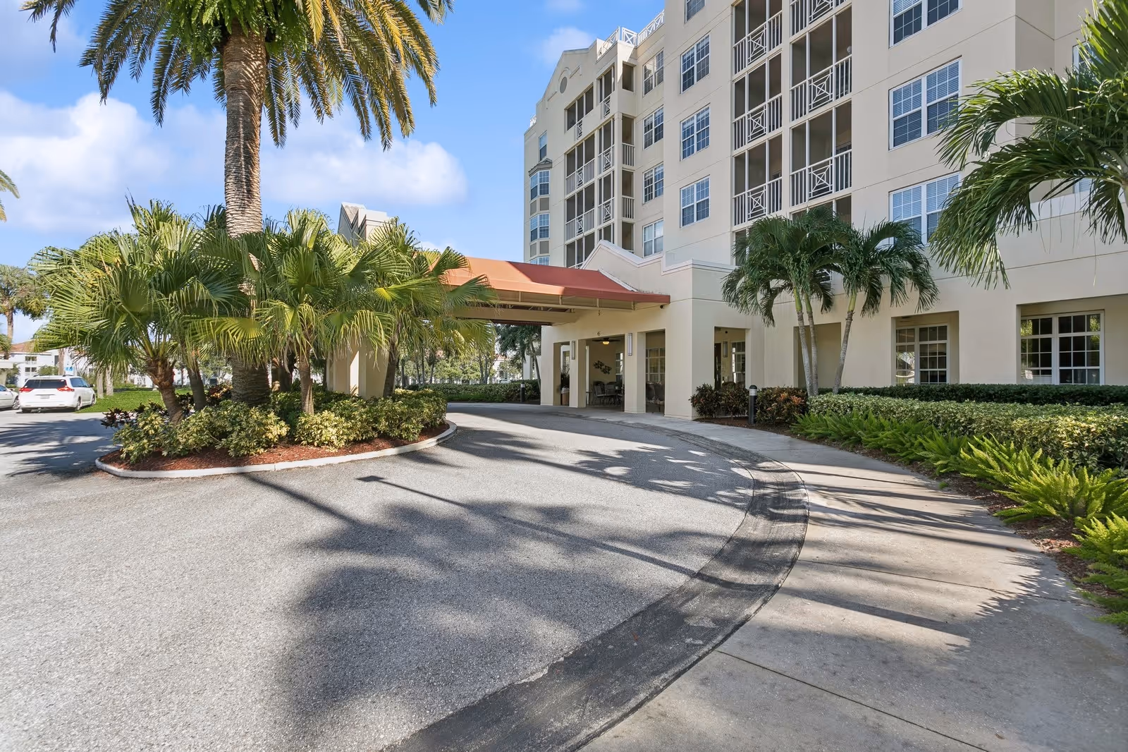 Exterior view of Stratford Court of Palm Harbor showing the entrance driveway with a covered drop-off area. The building is multi-story with balconies and large windows. There are palm trees and landscaped greenery around the driveway and building.