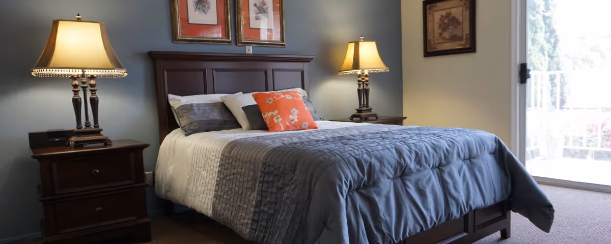 A neatly made bed with a gray and white striped comforter and multiple pillows, including an orange floral pillow, in a bedroom with two wooden nightstands each holding a lamp. There are framed pictures on the wall above the bed and a sliding glass door to the right letting in natural light.
