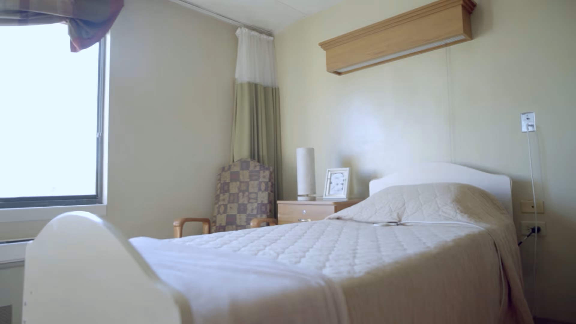 A simple bedroom in a senior living facility with a single bed covered in a beige quilted blanket, a wooden nightstand with a lamp and a framed picture, a patterned armchair, and a window with curtains allowing natural light to enter.