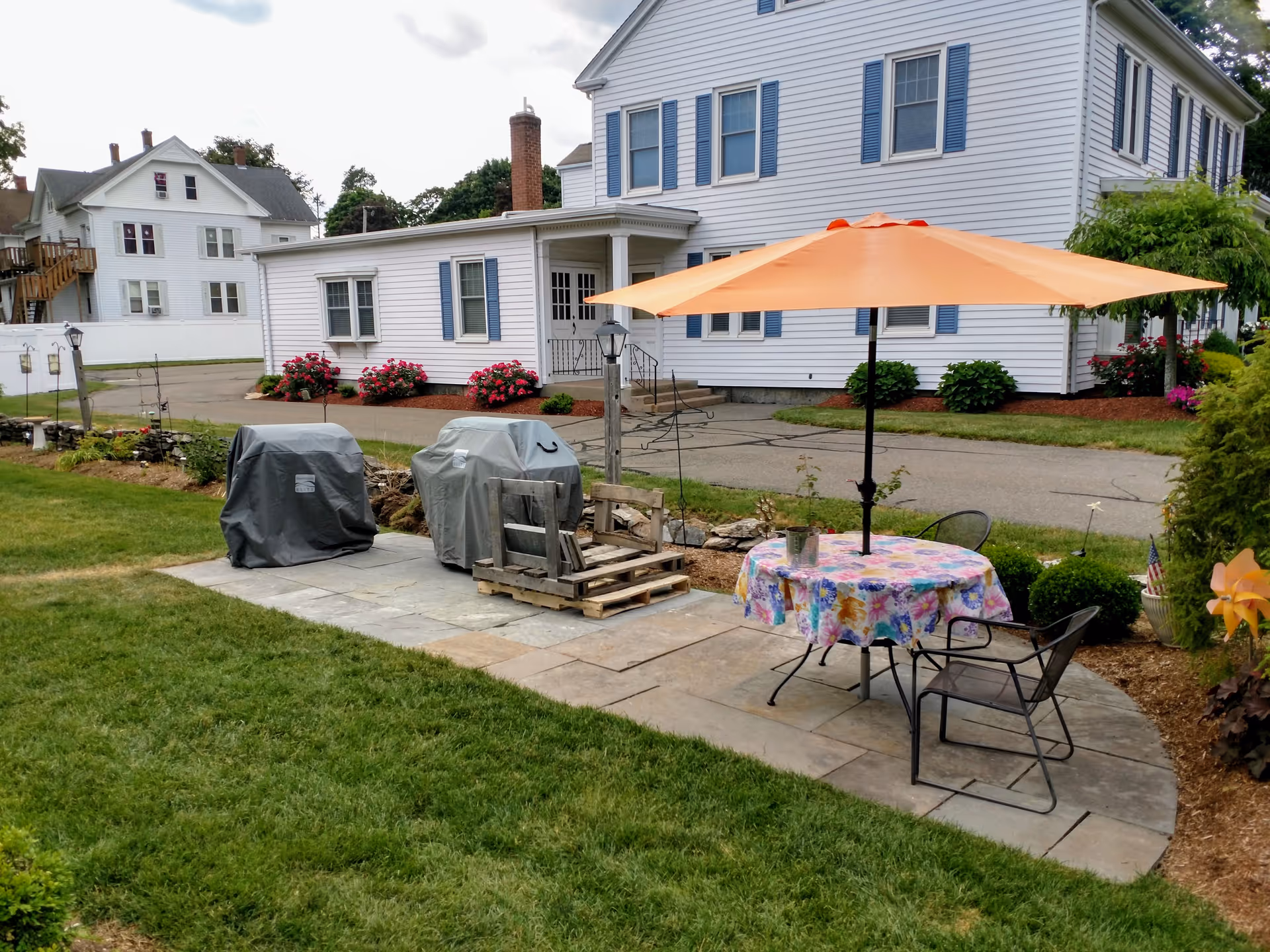Outdoor patio area with a round table covered by a colorful floral tablecloth and an orange umbrella. Two metal chairs are placed around the table. Nearby, two covered grills and a wooden pallet chair are on a stone-paved surface. The background shows a white building with blue shutters and flower beds with red flowers.