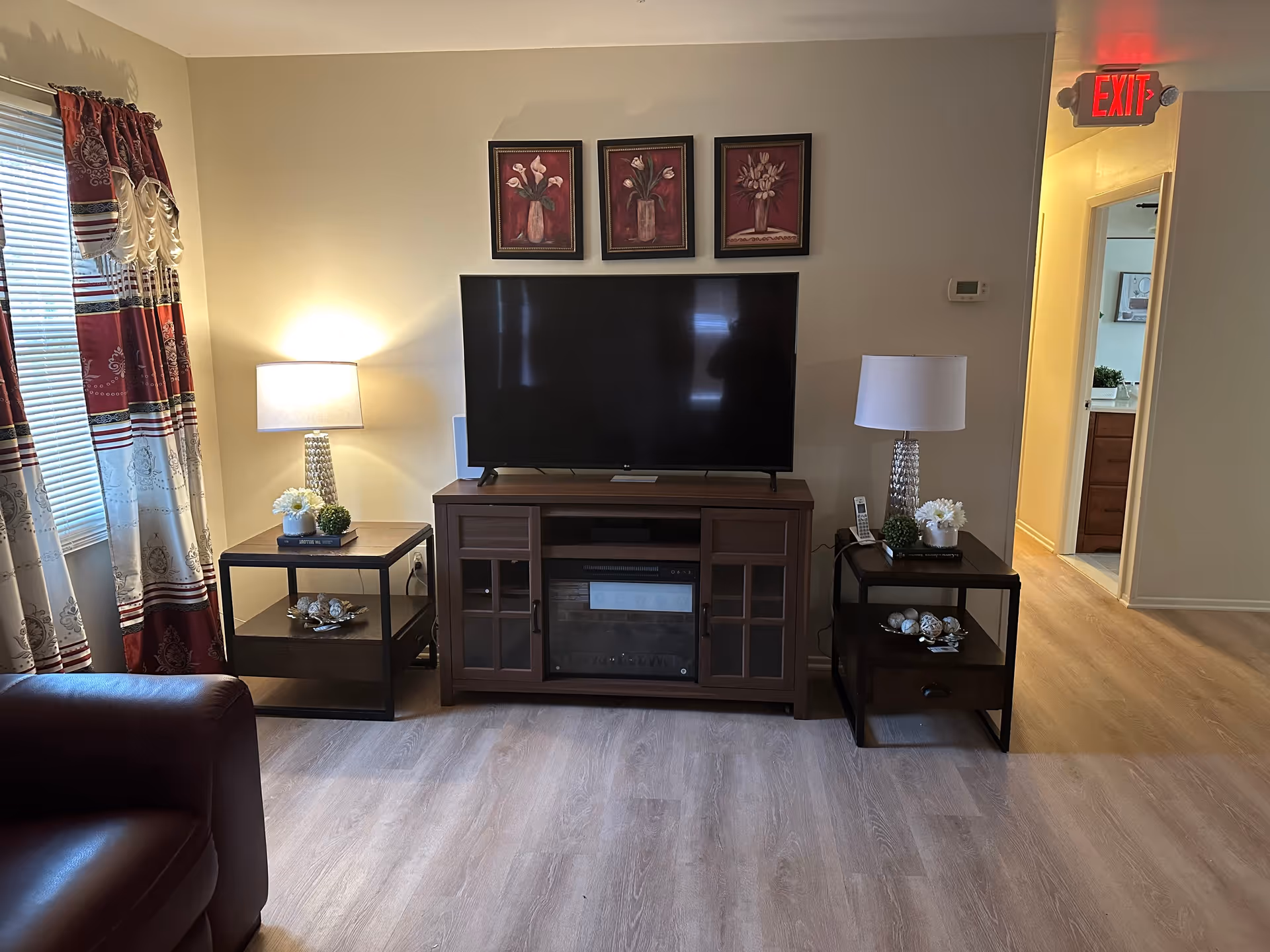 A cozy living room area with a flat-screen TV on a wooden TV stand that includes a built-in electric fireplace. On either side of the TV stand are matching dark wood side tables with table lamps, decorative plants, and ornaments. The room has light-colored walls and wood flooring. To the left, there is a window with red and white patterned curtains. An exit sign is visible above a doorway leading to another room.