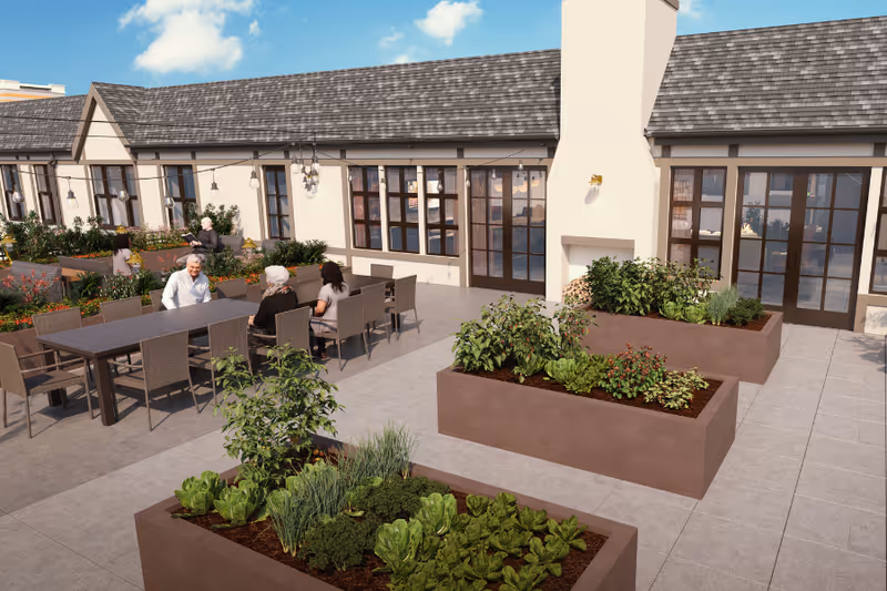 Outdoor patio area at Aegis Living Greenwood with raised garden beds containing various plants and vegetables. Several people are seated at a long table enjoying the space. The building has large windows and a chimney, with string lights hanging above the seating area under a blue sky with some clouds.