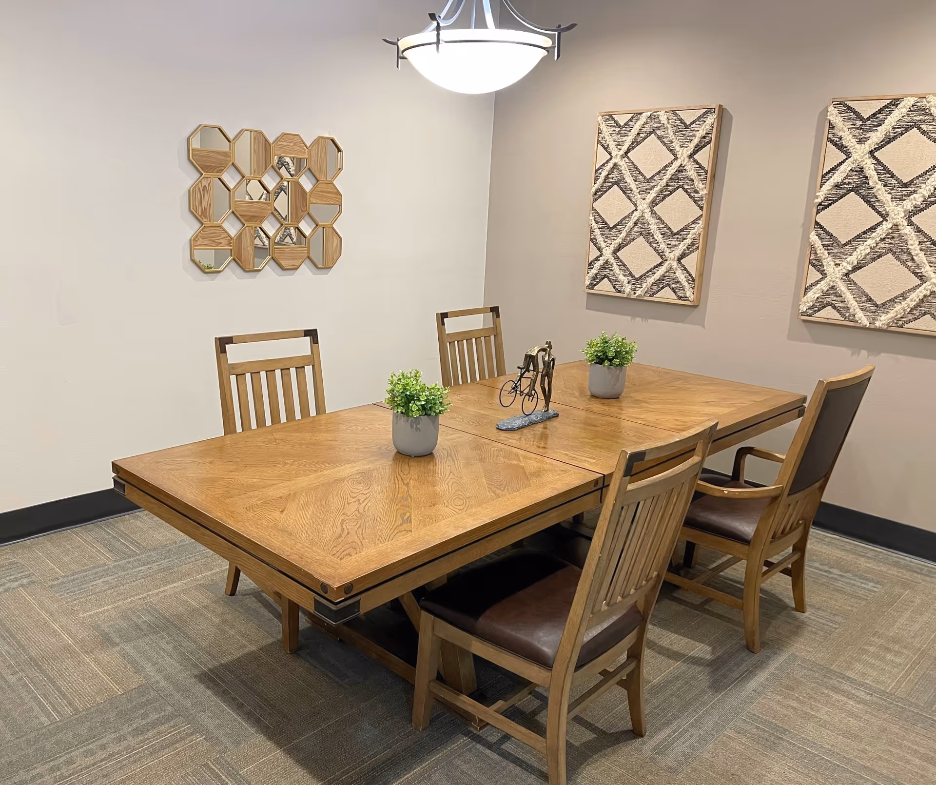 A dining room with a wooden table and four wooden chairs with brown cushions. The table has two small potted plants and a small decorative sculpture of a person riding a bicycle. The walls are beige with two patterned wall hangings and a decorative mirror with multiple hexagonal sections. A ceiling light fixture hangs above the table.