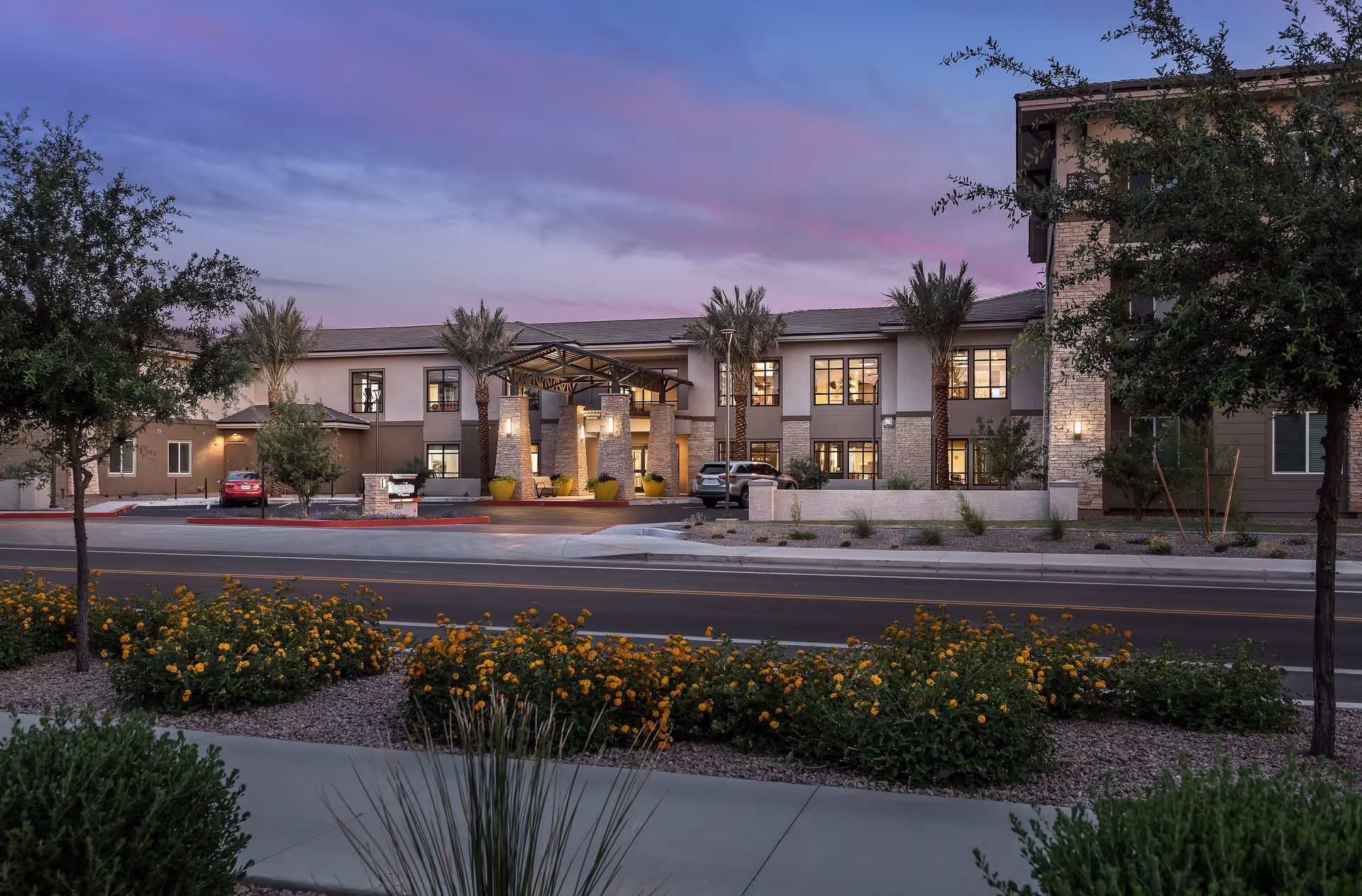 Two-story senior living building front with illuminated entrance, palm trees and landscaped beds at dusk.