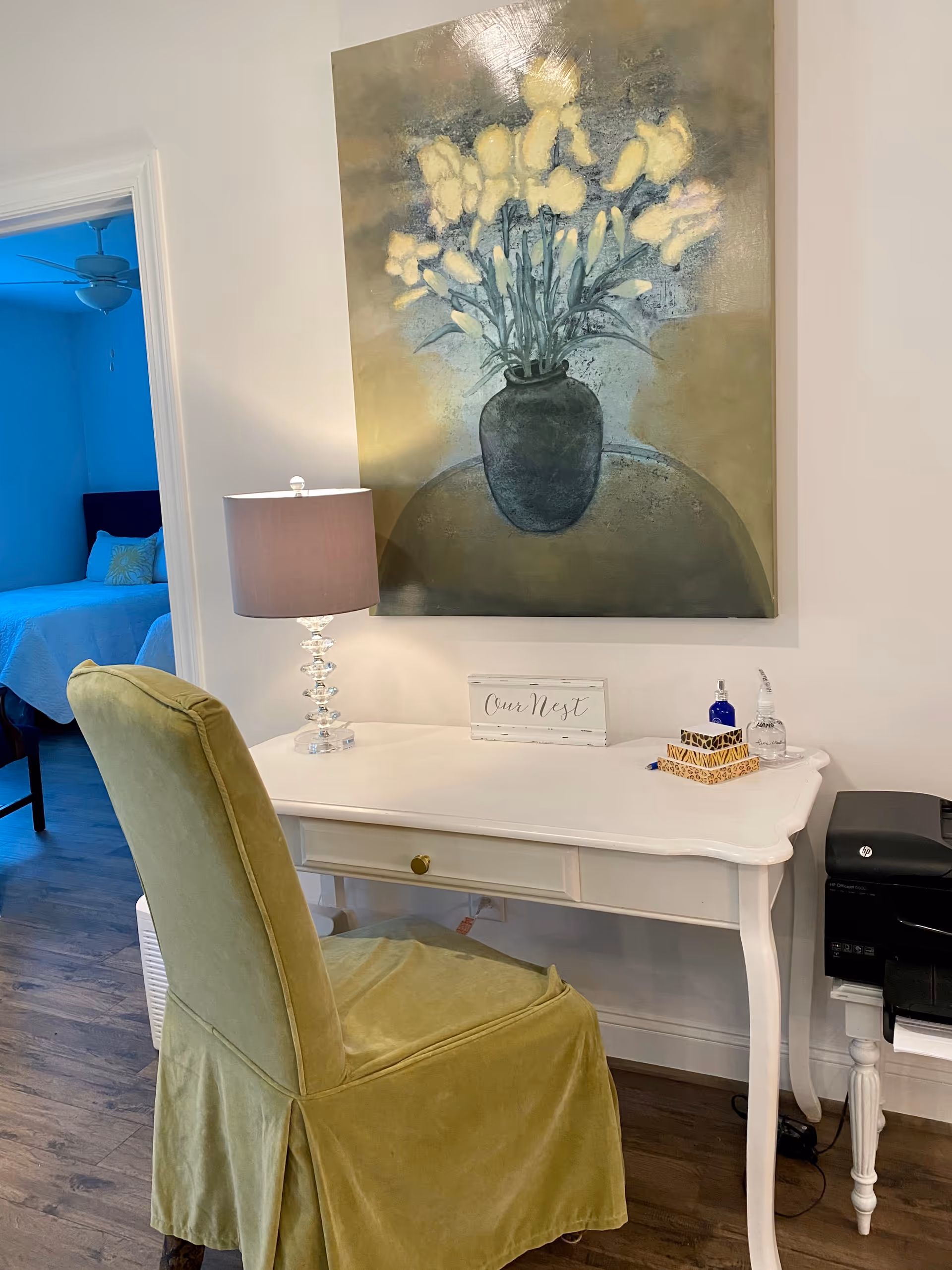 A cozy interior corner featuring a white desk with a drawer, a green upholstered chair, a table lamp with a gray shade, a decorative sign that reads 'Our Nest', two small bottles, and a box of tissues. Above the desk hangs a large painting of a vase with yellow flowers. In the background, a bedroom with a blue bedspread and a ceiling fan is partially visible.