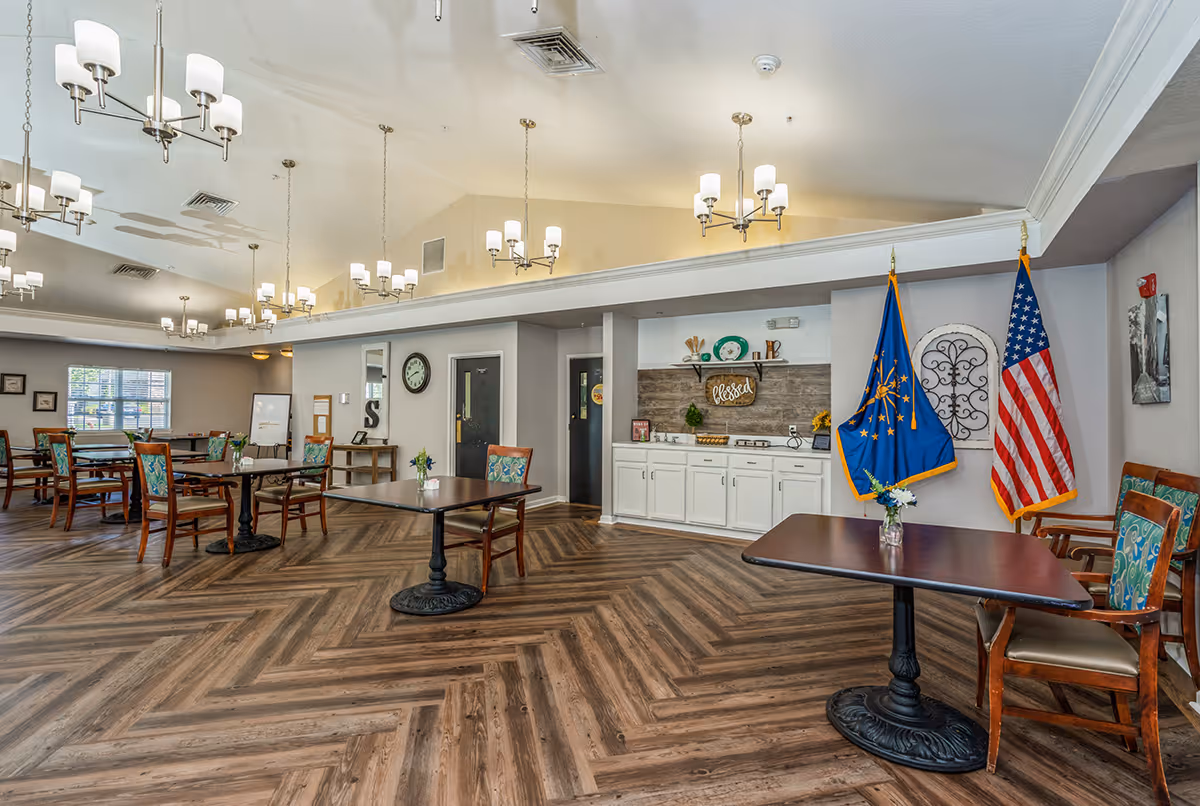 A spacious dining room with several wooden tables and chairs arranged neatly. The floor has a herringbone wood pattern, and multiple modern chandeliers hang from the ceiling. On the right side, there is a small counter with white cabinets and decorative items, including a sign that says 'blessed'. Two flags, one of Indiana and one American flag, stand next to the counter. The room has large windows allowing natural light to enter.