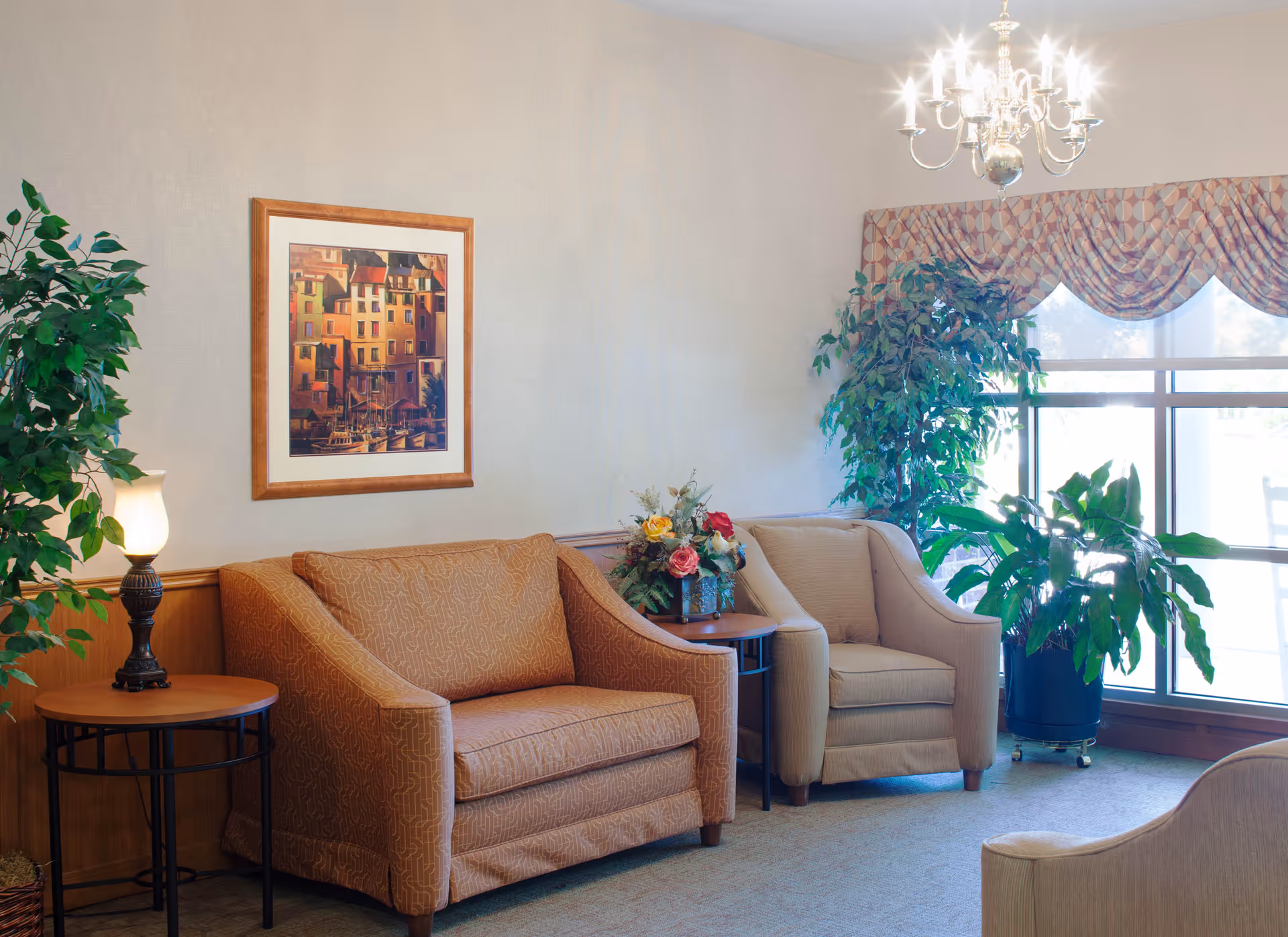 A cozy sitting area with two upholstered armchairs and a small round wooden table with a lamp. There are two large potted plants near a window with patterned valance curtains. A framed painting of colorful buildings hangs on the wall above the chairs, and a chandelier is visible on the ceiling.