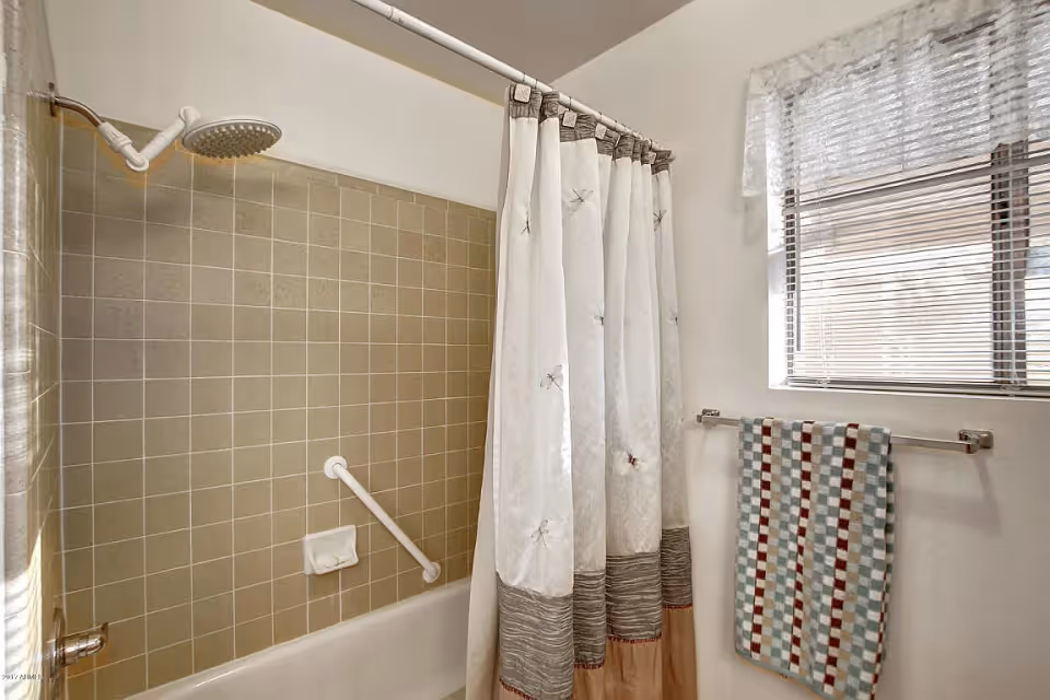 Bathtub and shower with beige tiled wall, white shower curtain and grab bar, a window with blinds, and a patterned towel on a wall-mounted rack.