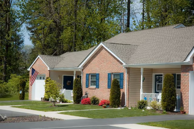 Single-story residential building with brick and siding exterior, blue shutters, small front porches, manicured lawns, shrubs, and an American flag displayed near the garage. Trees and greenery are visible in the background under a clear sky.