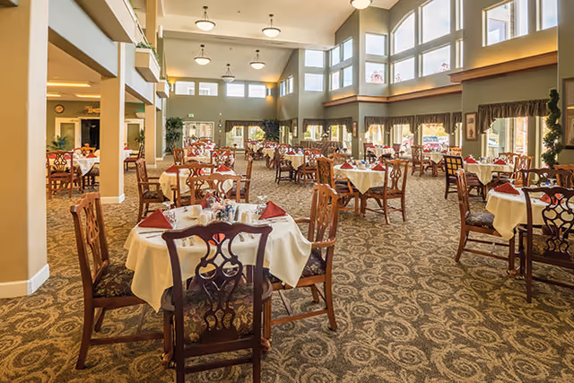 Spacious dining room with round tables dressed in white tablecloths and red napkins under high ceilings and large windows.