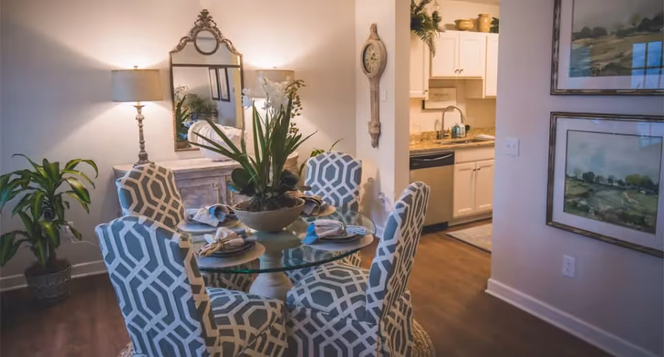 A dining area with a round glass table set for four, surrounded by patterned upholstered chairs. A large decorative plant is in the center of the table. Behind the table is a sideboard with a lamp and a large ornate mirror. To the right, there is a partial view of a kitchen with white cabinets and a granite countertop. The walls are decorated with framed artwork and a wall clock.