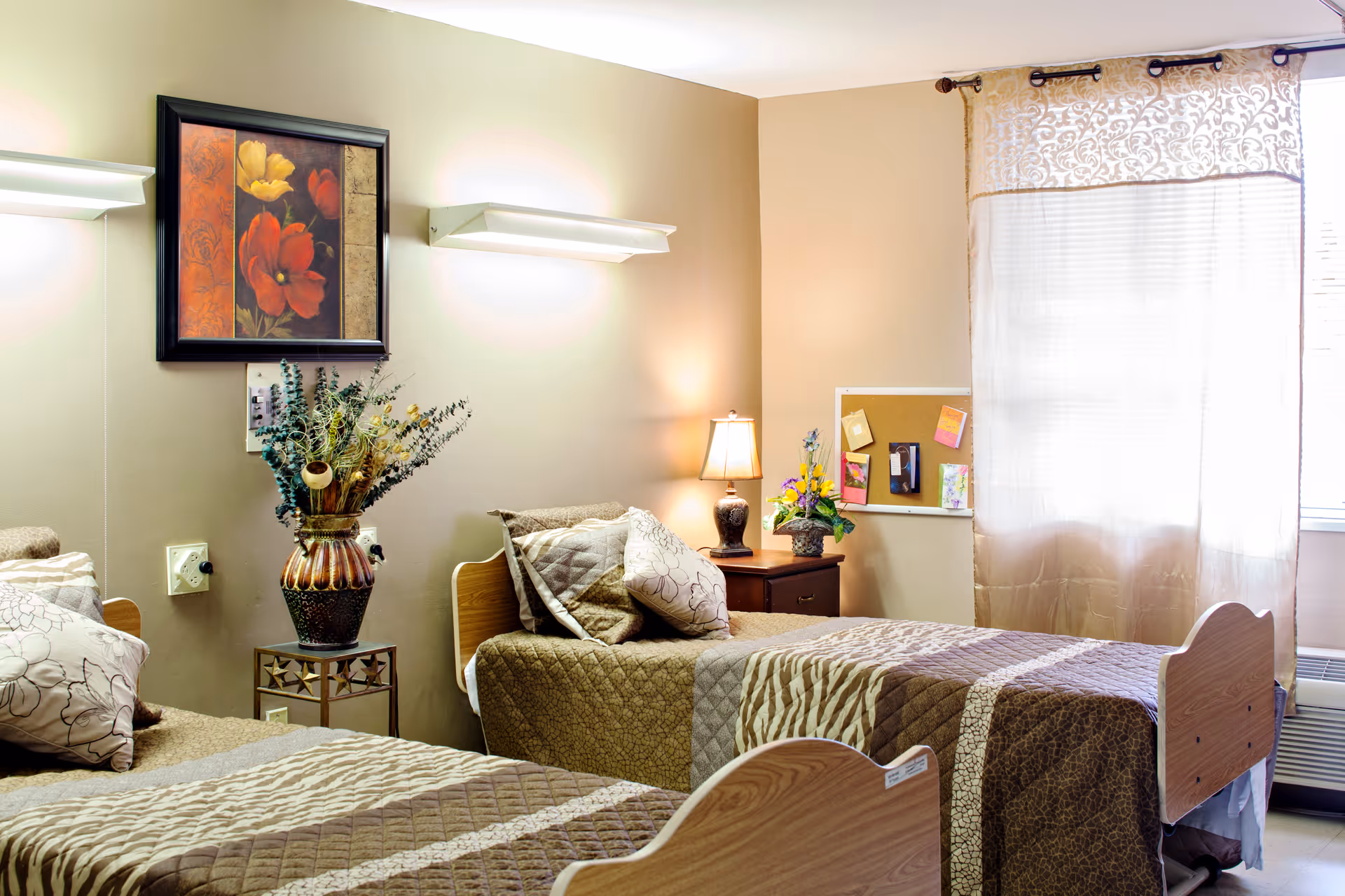 A cozy room with two single beds featuring brown and beige patterned bedspreads and pillows. Between the beds is a small table with a decorative vase holding dried flowers. On the wall above the table is a framed floral painting. A nightstand with a lamp and a small flower arrangement is next to one of the beds. A window with sheer curtains allows natural light into the room, and a bulletin board with pinned notes is visible on the wall.