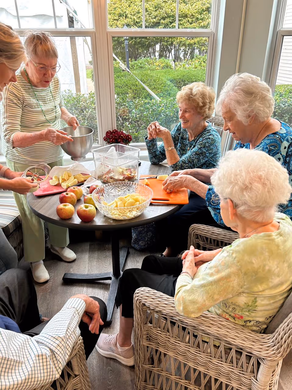 A group of elderly people sitting around a round table near large windows, peeling and cutting apples. There are several apples and a bowl of sliced apples on the table. The room is bright with natural light and has a cozy atmosphere.